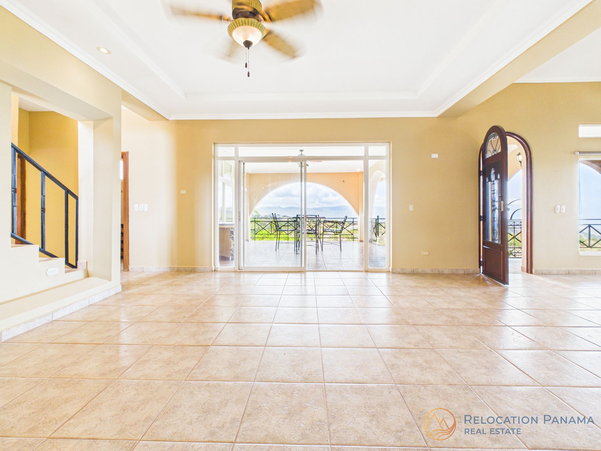 Una habitación luminosa y vacía con suelo de baldosas color canela, paredes color crema, un ventilador de techo, una escalera a la izquierda y vistas a un patio al fondo.