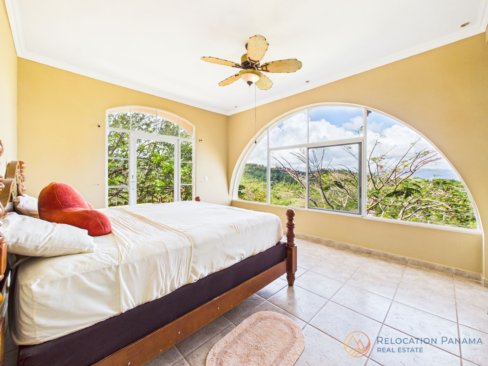 Un dormitorio espacioso con cama, paredes de color amarillo claro, suelo de baldosas y un gran ventanal arqueado con vistas panorámicas al exterior.