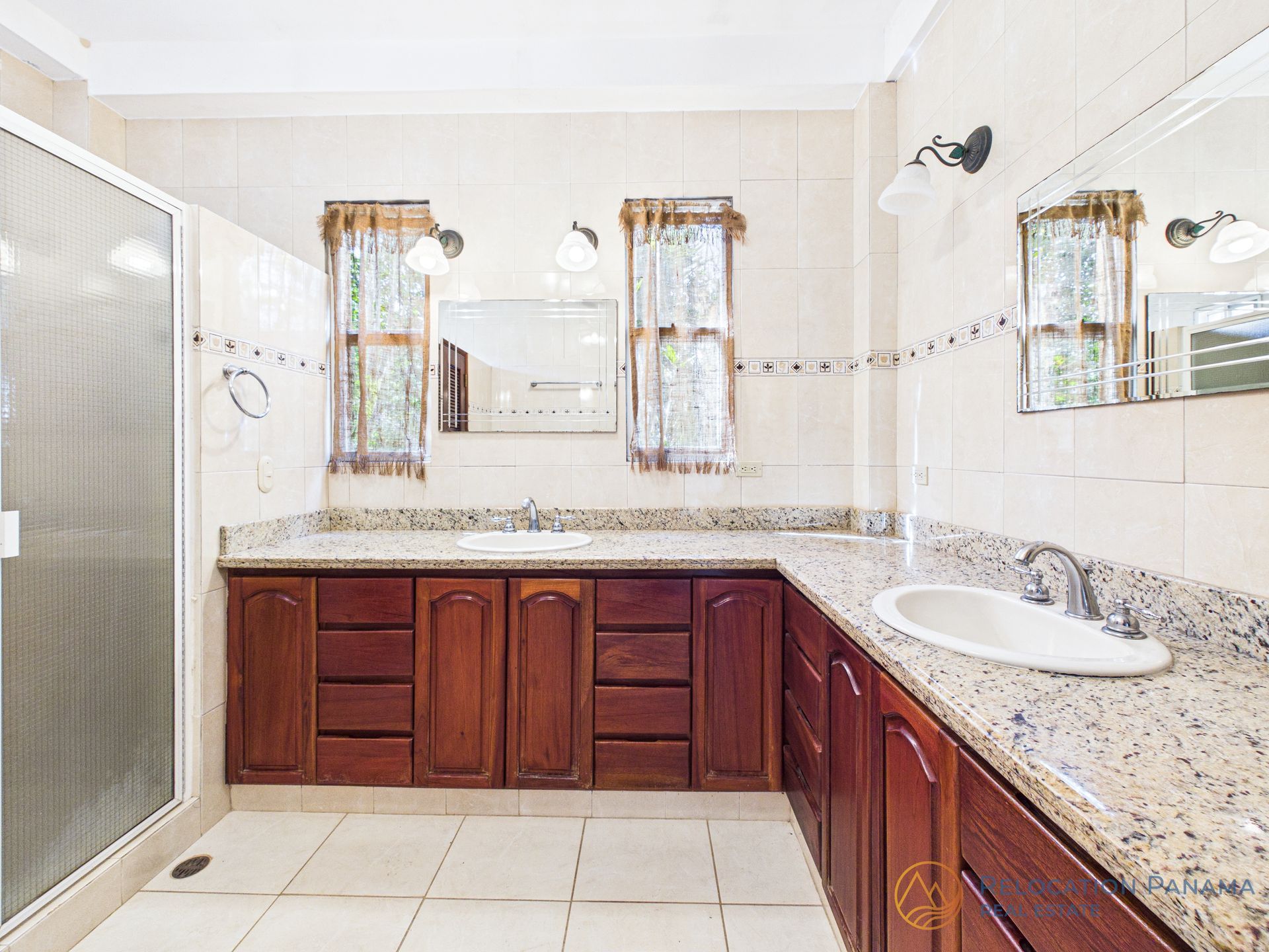 Un baño con tocador doble de madera con encimeras de granito, paredes alicatadas, ducha de cristal y dos ventanas.