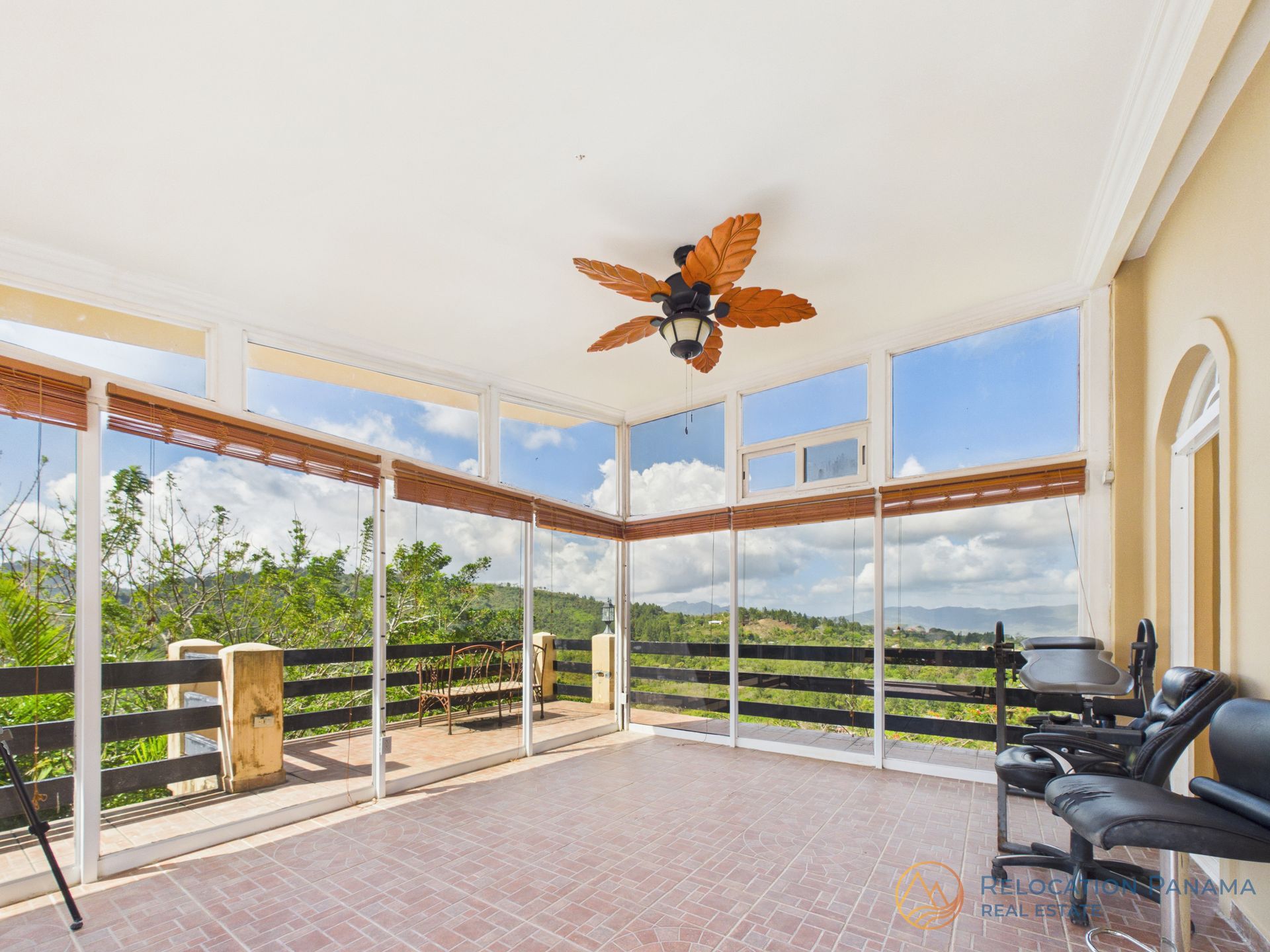 Una terraza acristalada con suelo de baldosas, un ventilador de techo de madera y grandes ventanales que ofrecen una vista panorámica de las colinas onduladas y el cielo.