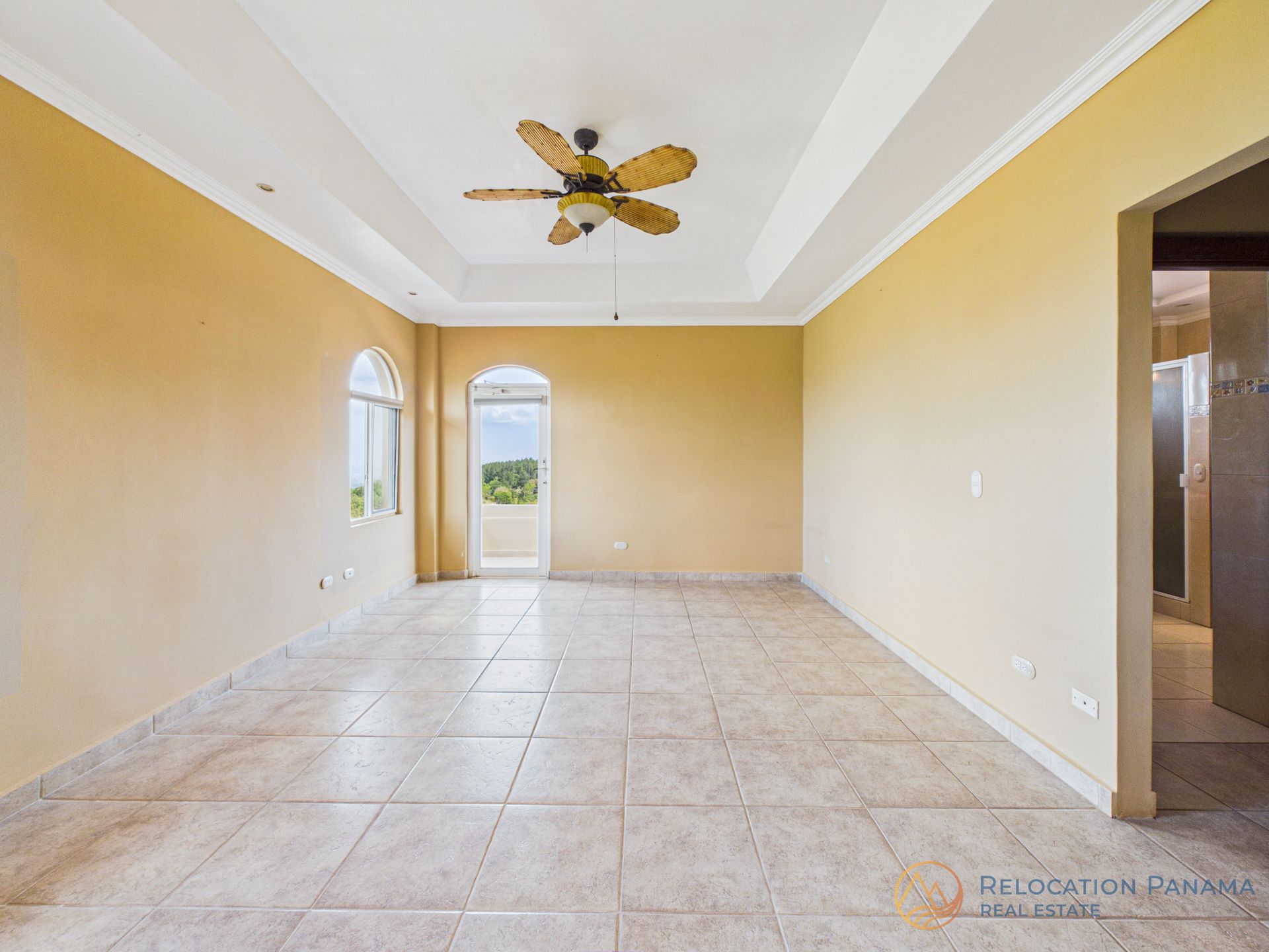 Una habitación luminosa y vacía con paredes de color beige, suelo de baldosas claras, un techo artesonado con ventilador y una puerta y ventana arqueadas.
