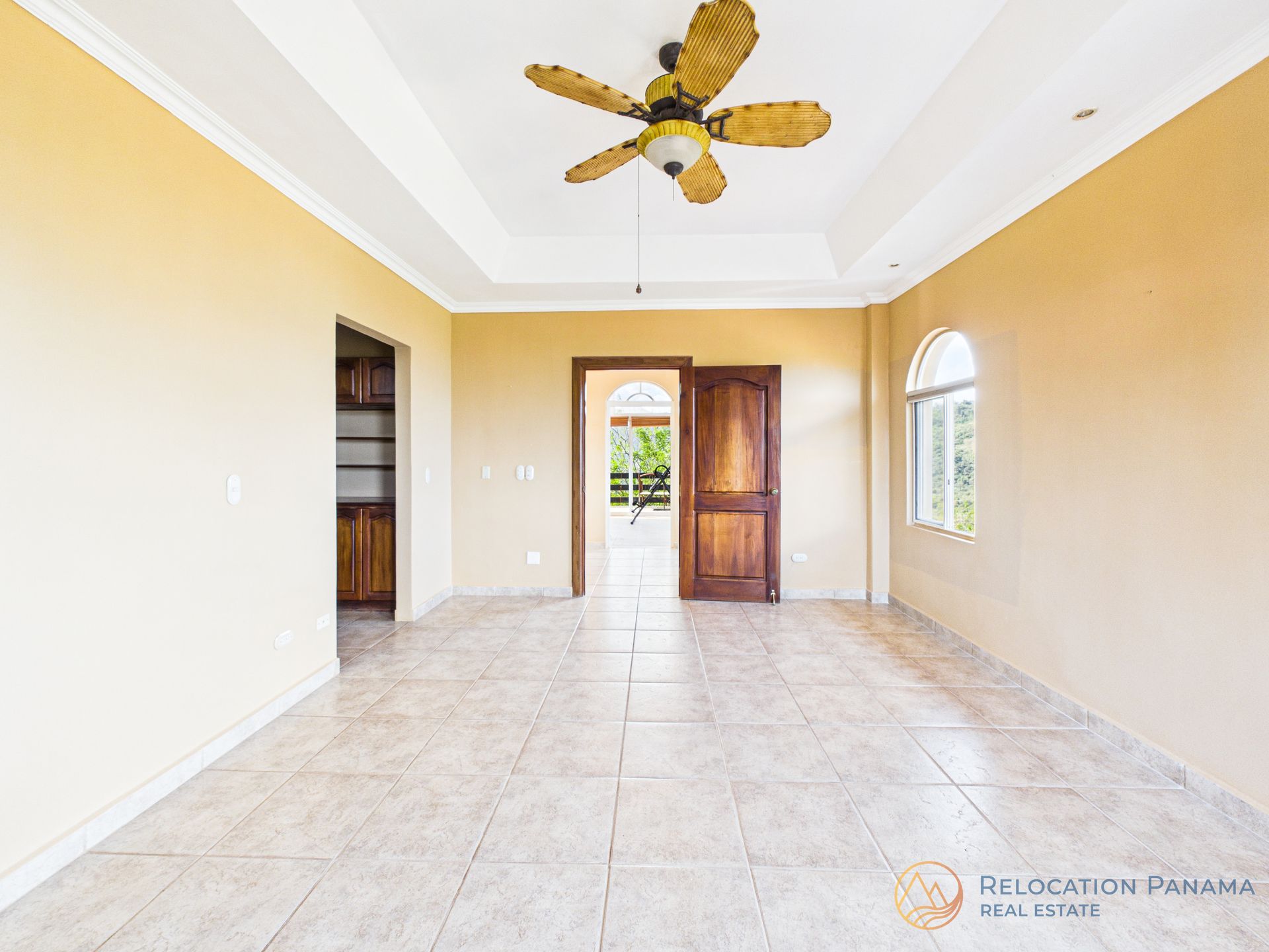 Una habitación vacía y soleada con paredes de color beige, suelos de baldosas, un ventilador de techo central y puertas abiertas que dan a otras habitaciones.