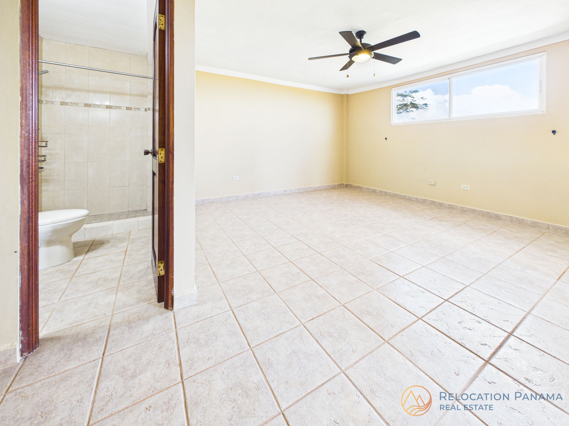 Un dormitorio vacío con paredes color crema, suelo de baldosas, un ventilador de techo y una puerta abierta que da a un baño contiguo.