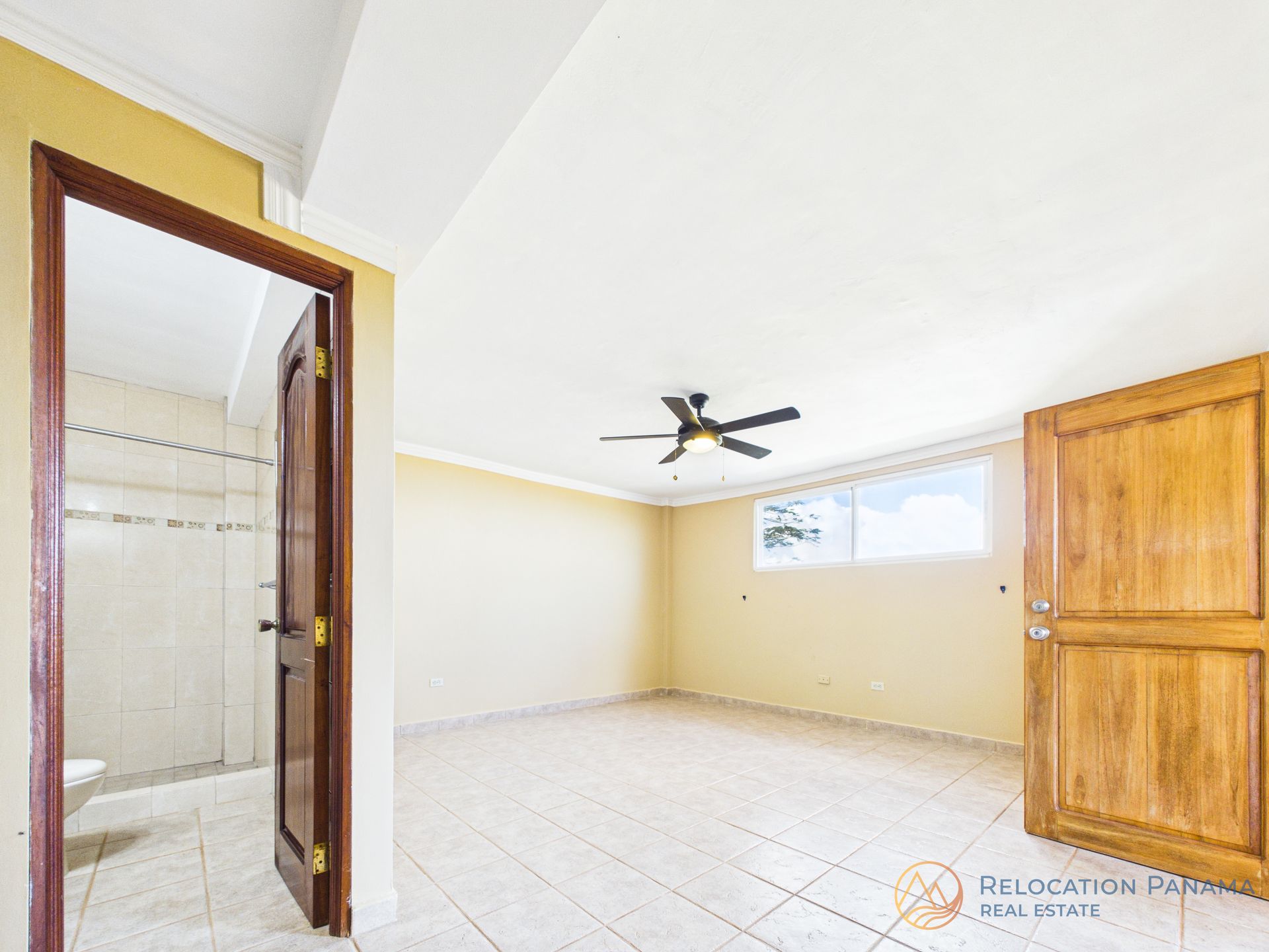 Una habitación luminosa y vacía con suelo de baldosas de color claro, paredes amarillas, un ventilador de techo y una puerta de madera abierta que da a un baño.