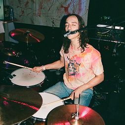 Drummer with long hair, wearing tie-dye shirt, playing drums with closed eyes and mic at his mouth.