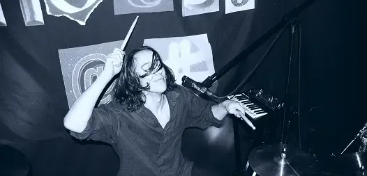 A musician playing drums with intense focus, dark clothing, stage setting.