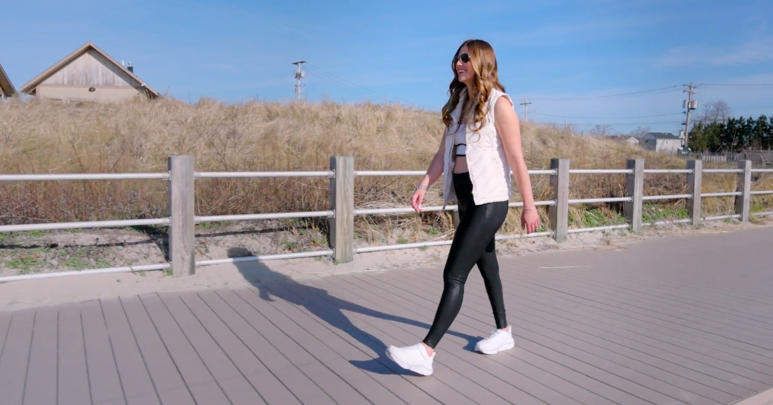 Alexa is walking down a boardwalk next to a fence on the beach wearing white cadense adaptive sneakers