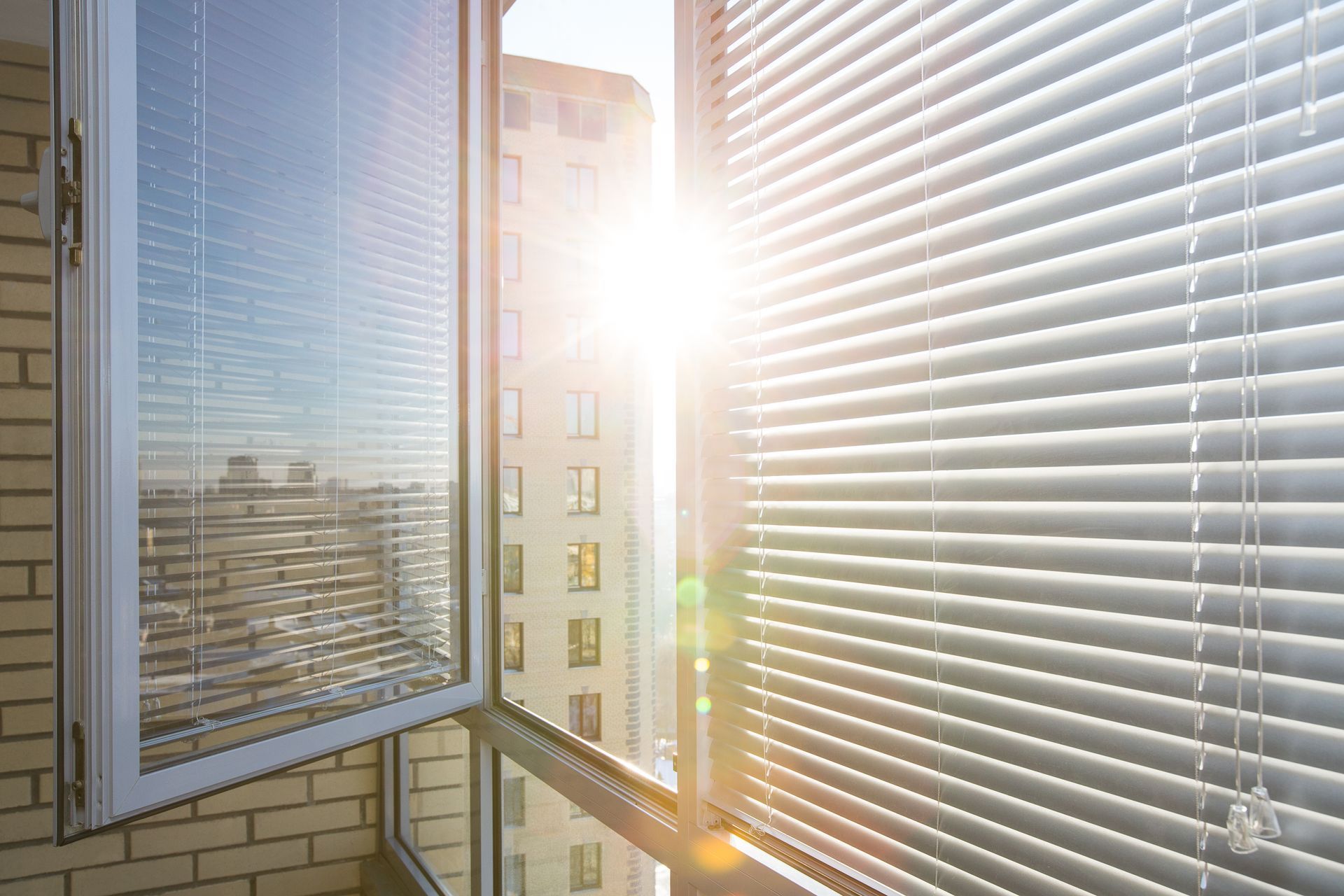 Sunlit balcony with blinds, brick wall, and bright glare over a city view