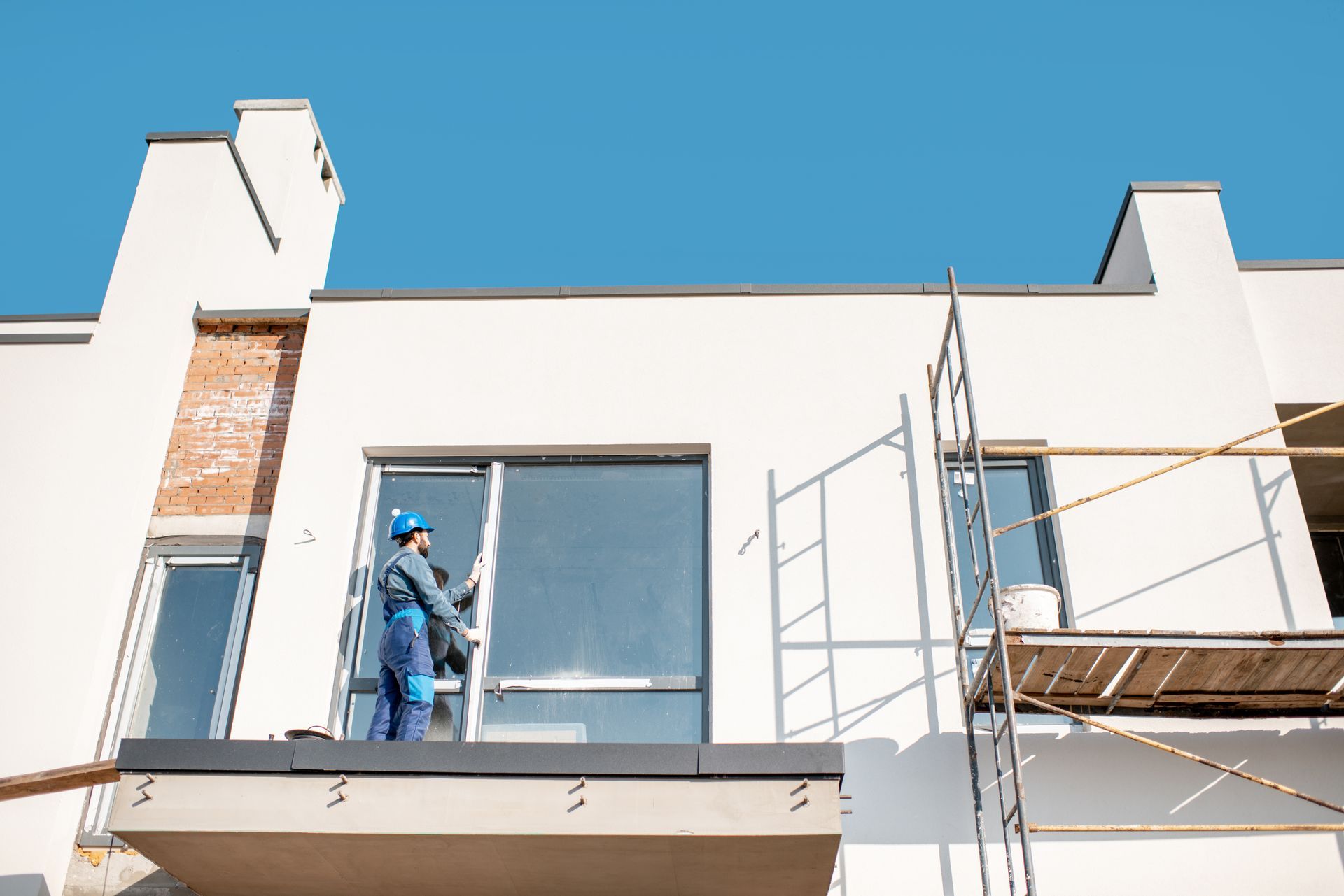 Worker on scaffolding painting a white building facade under a blue sky