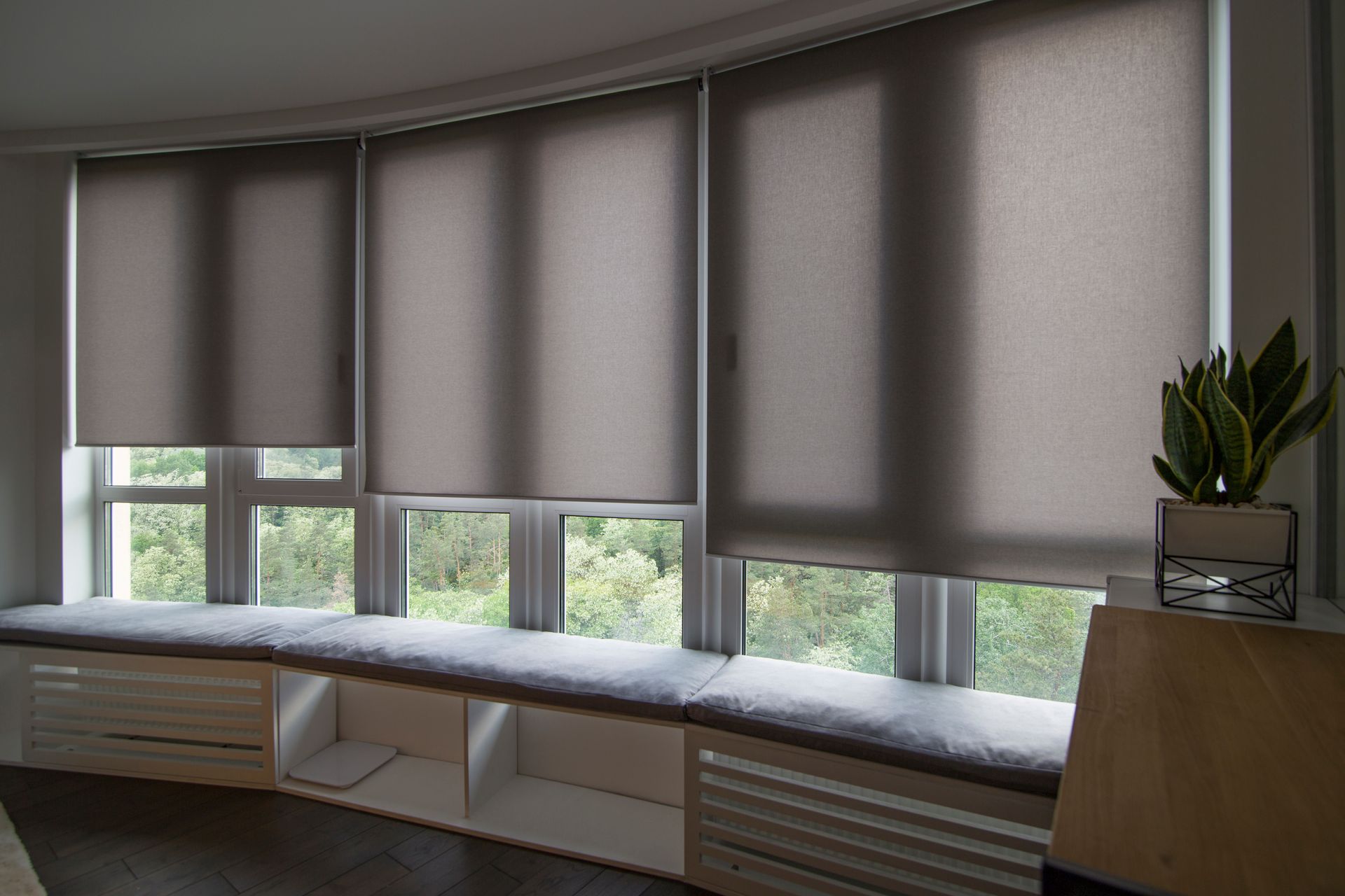 Curved window seat with gray blinds, built-in benches, and potted plants in a bright room