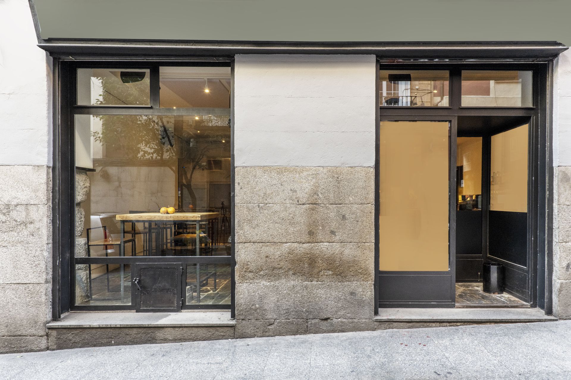 Two storefront windows on a gray facade, one with a table visible inside and the other with a tan door.