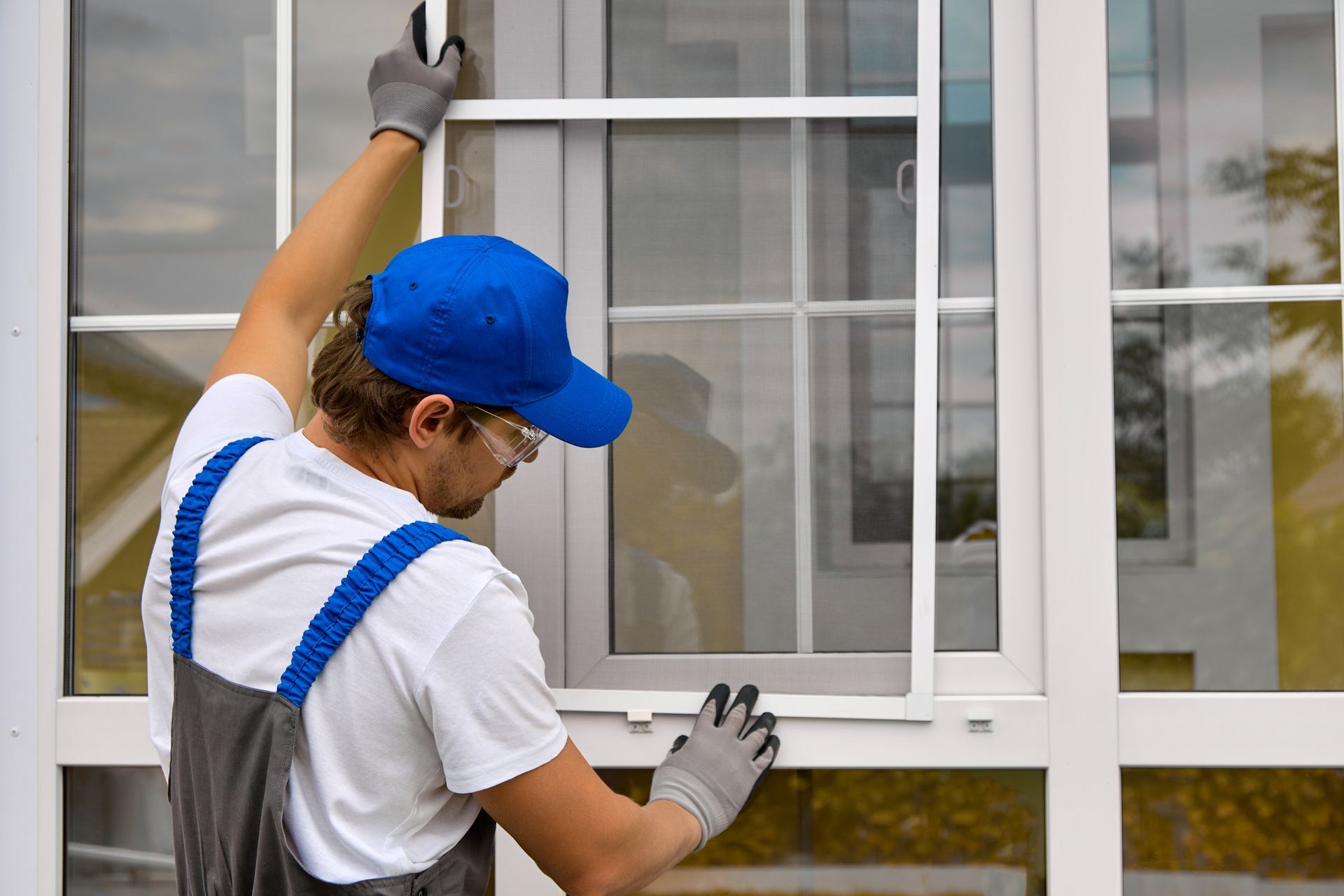 Window cleaner in blue cap and gloves washing a large white-framed window outdoors