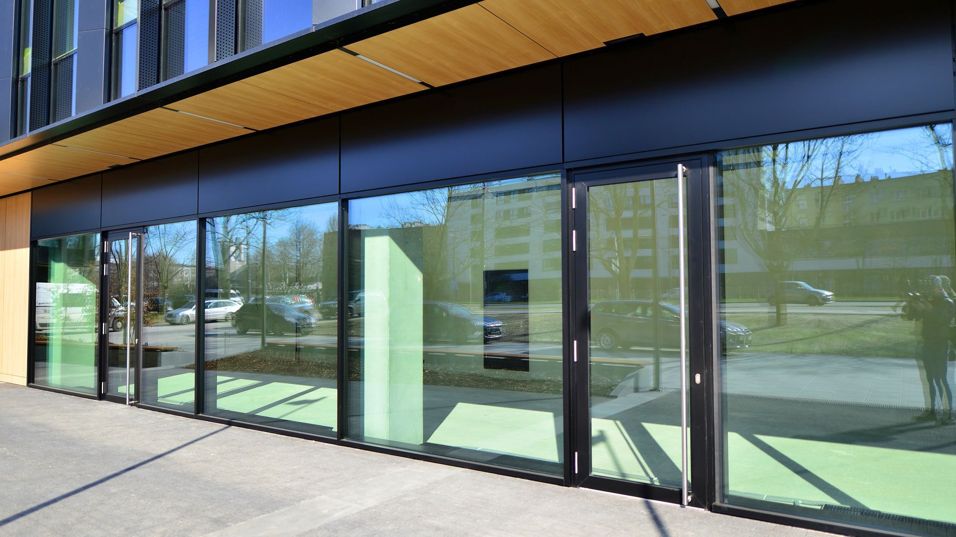 Modern glass storefront with black frames and green-tinted windows, reflecting trees and a parking lot