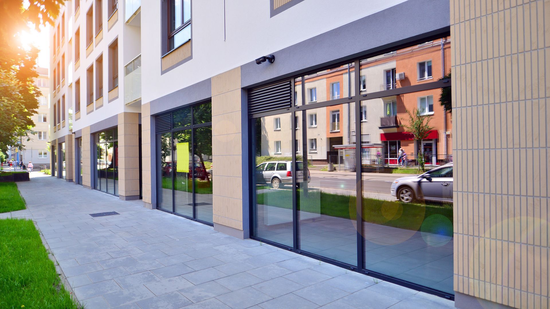 Modern glass-fronted building entrance along a city sidewalk, with reflections of nearby street and buildings