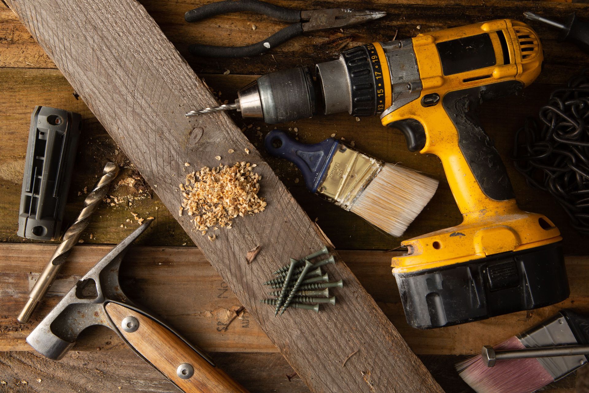 Yellow cordless drill, hammer, saw, and screws on a wooden workbench with wood shavings