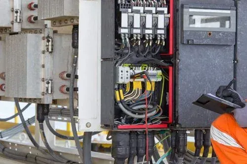 An electrician in orange coveralls inspects a control panel with wires and cables, using a tablet.