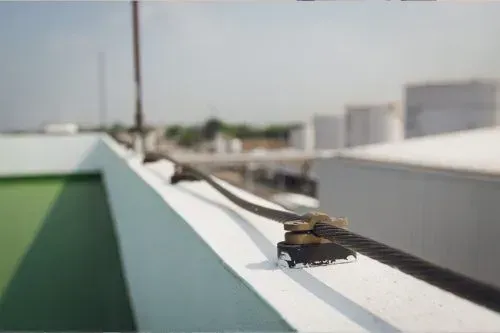 Steel cable running along the edge of a white rooftop, secured by metal clamps, with a blurred background.