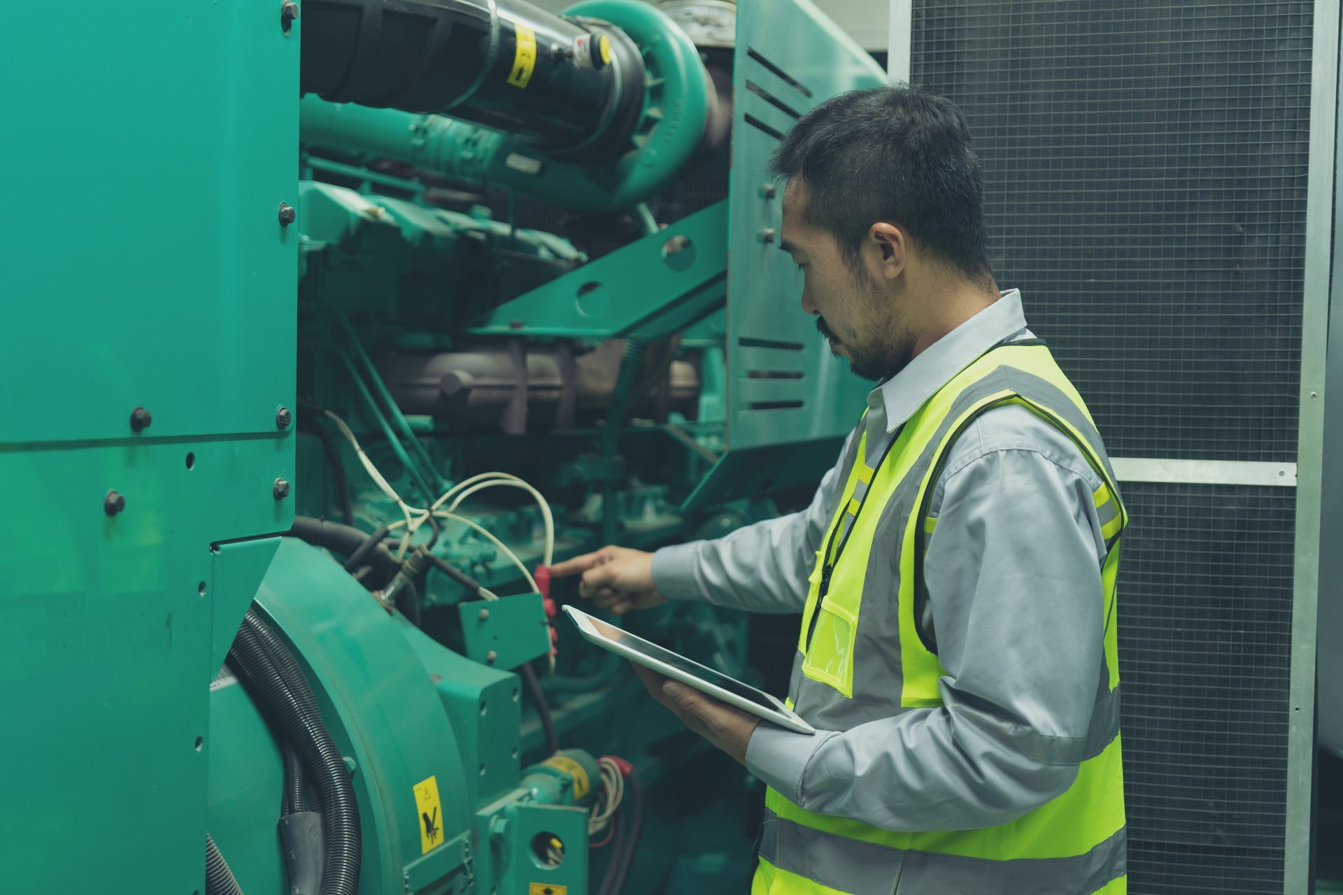 Man in safety vest inspects green industrial machine with tablet.