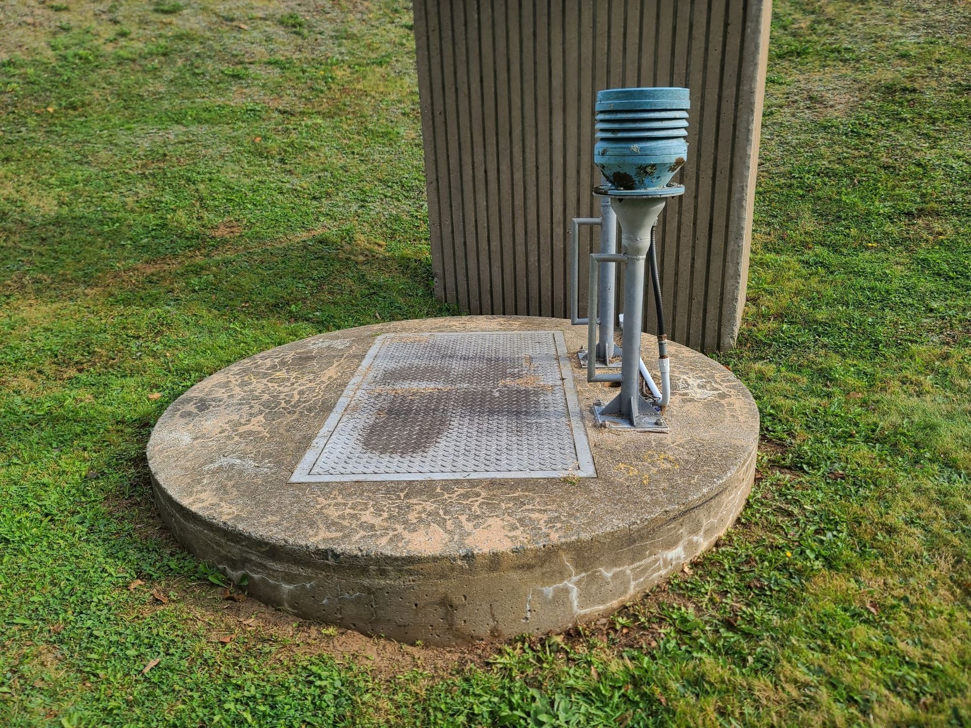 Concrete well with metal grate and weather monitoring equipment on green grass.