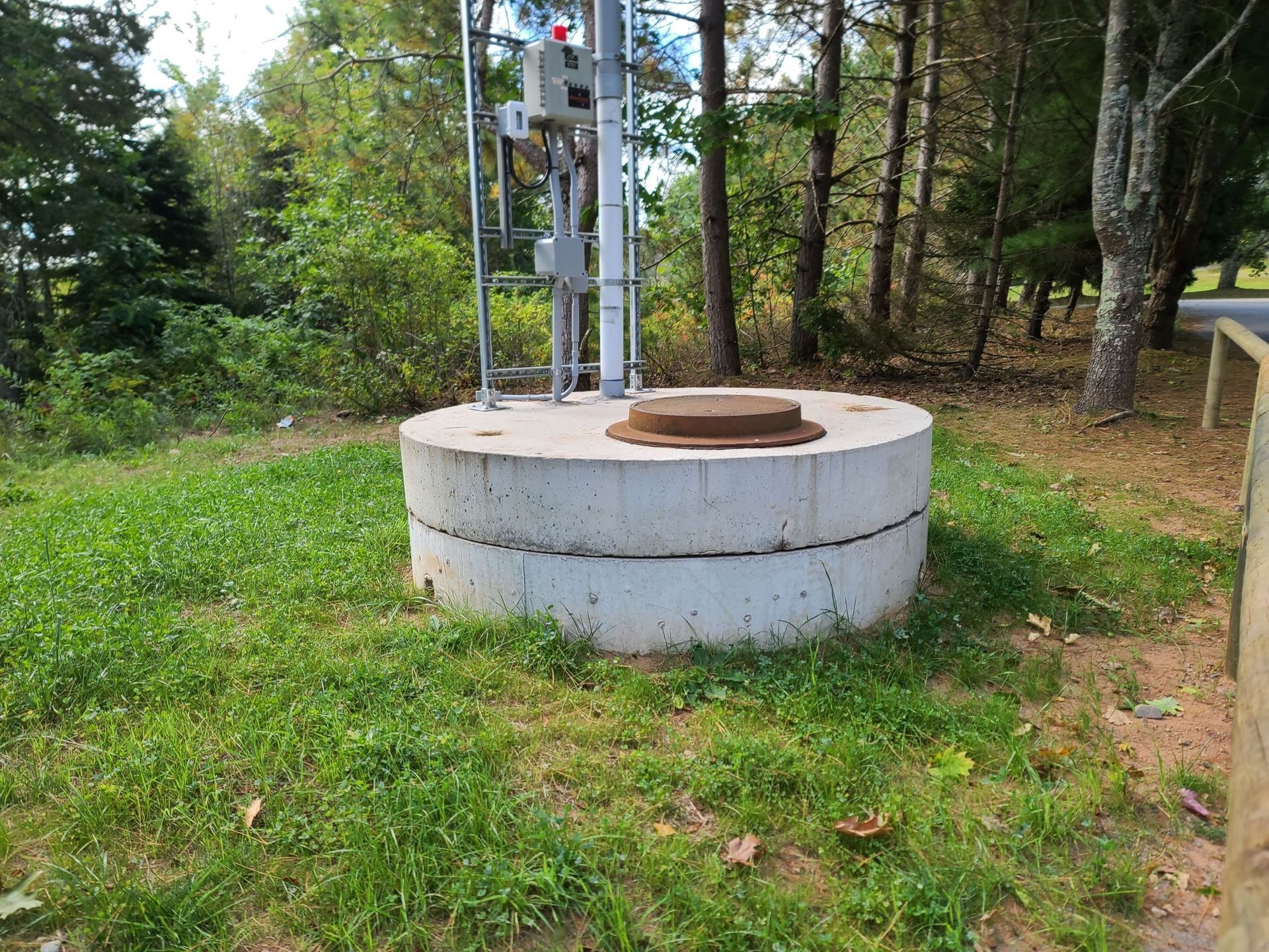 Concrete cylinder base with metal hatch and tower, surrounded by green grass, trees, and a wooden fence.