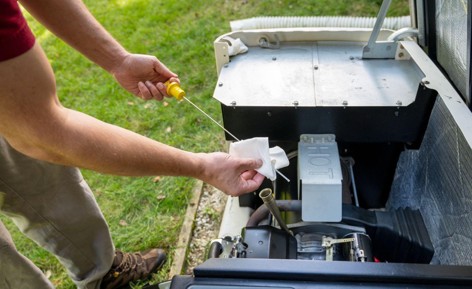 Person checking the oil level of a generator with a dipstick, wiping it with a white cloth outdoors on grass.