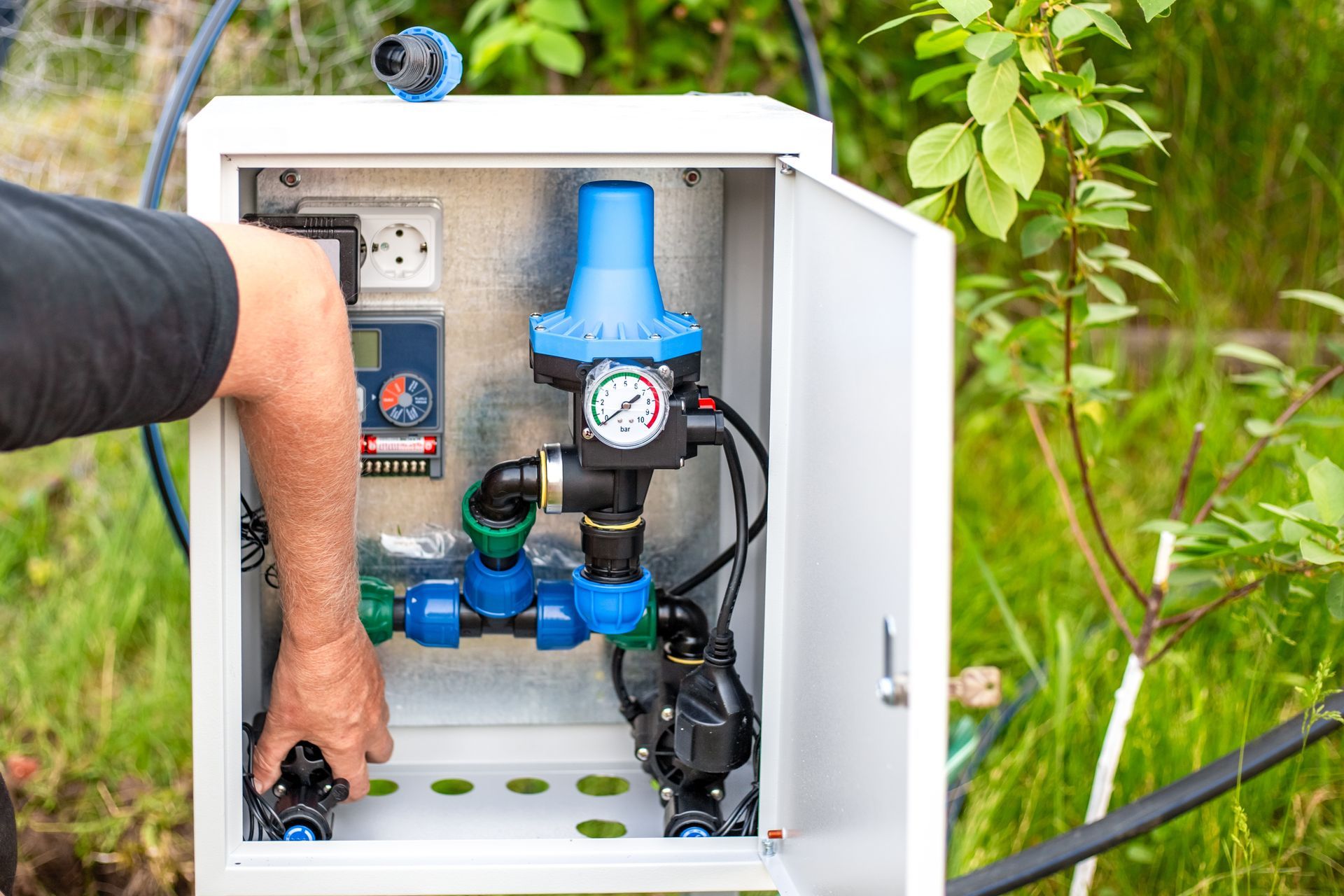 Person working on a white water pump control box in a garden setting with a blue pressure regulator.