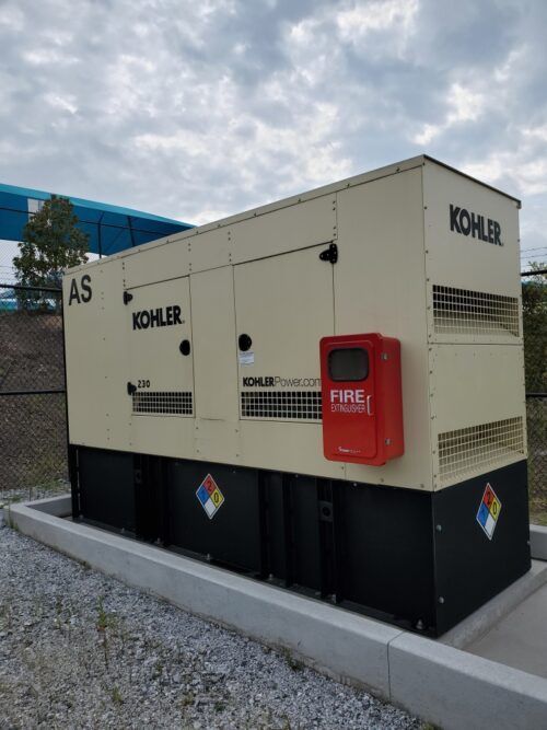 Large beige and black Kohler generator box outdoors with a red fire extinguisher cabinet.