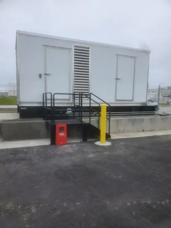 White industrial building with black stairs, handrails, and a yellow bollard on concrete. A red fire extinguisher case is nearby.