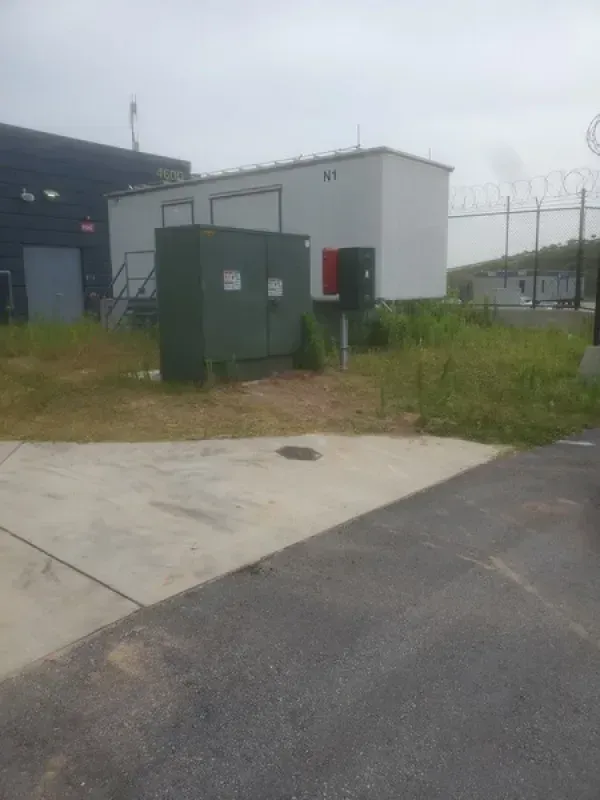 Green electrical box, white building, chain-link fence with razor wire, and asphalt in an outdoor setting.