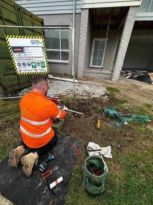 A Man is Kneeling Down in Front of a House Working on a Pipe — JMB Electrical Services In Port Kembla, NSW