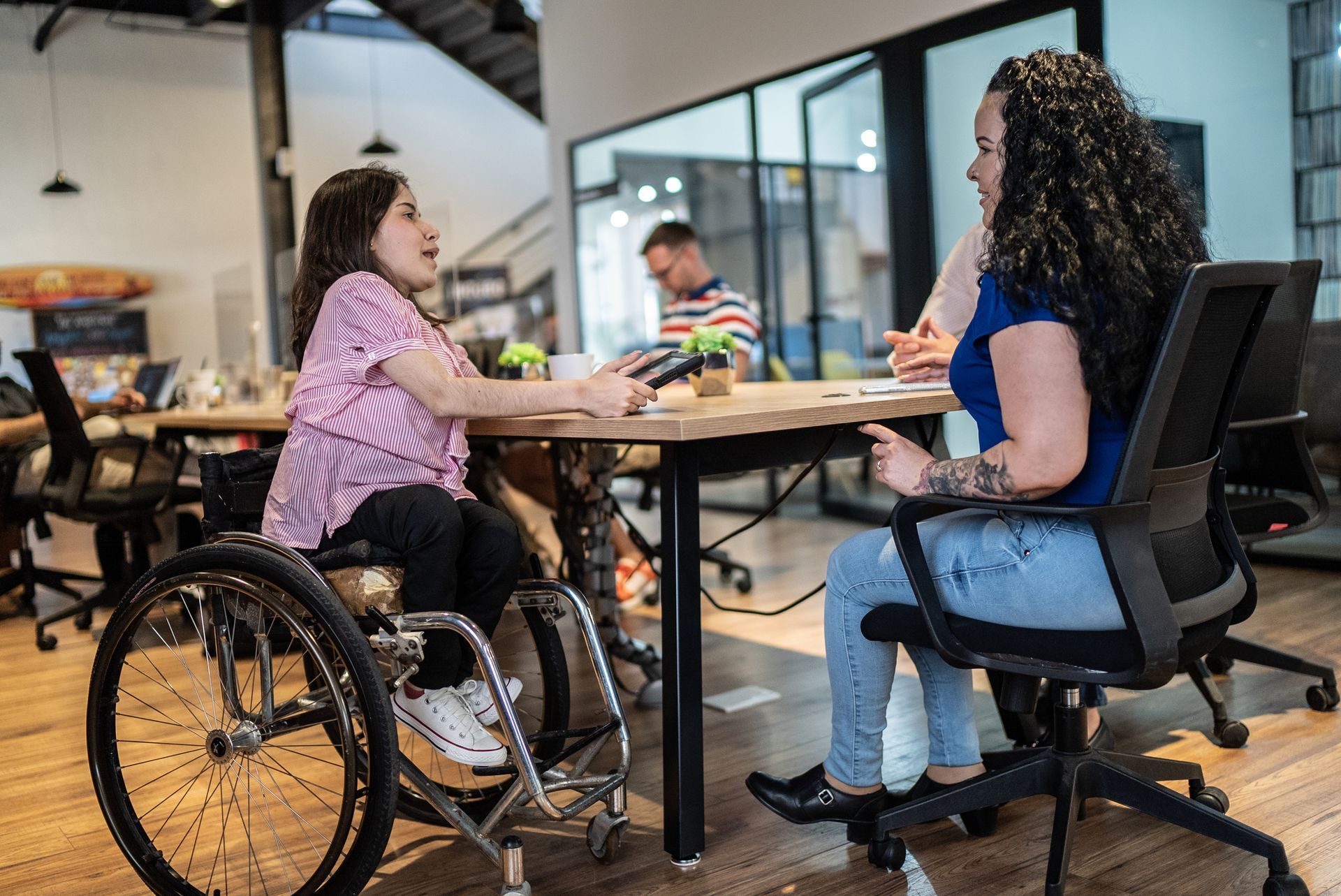 A woman in a wheelchair is sitting at a table talking to another woman.