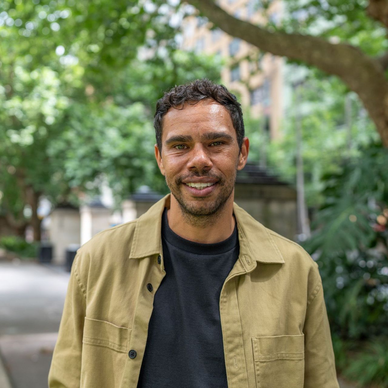 A man wearing a tan jacket and a black shirt is smiling for the camera.