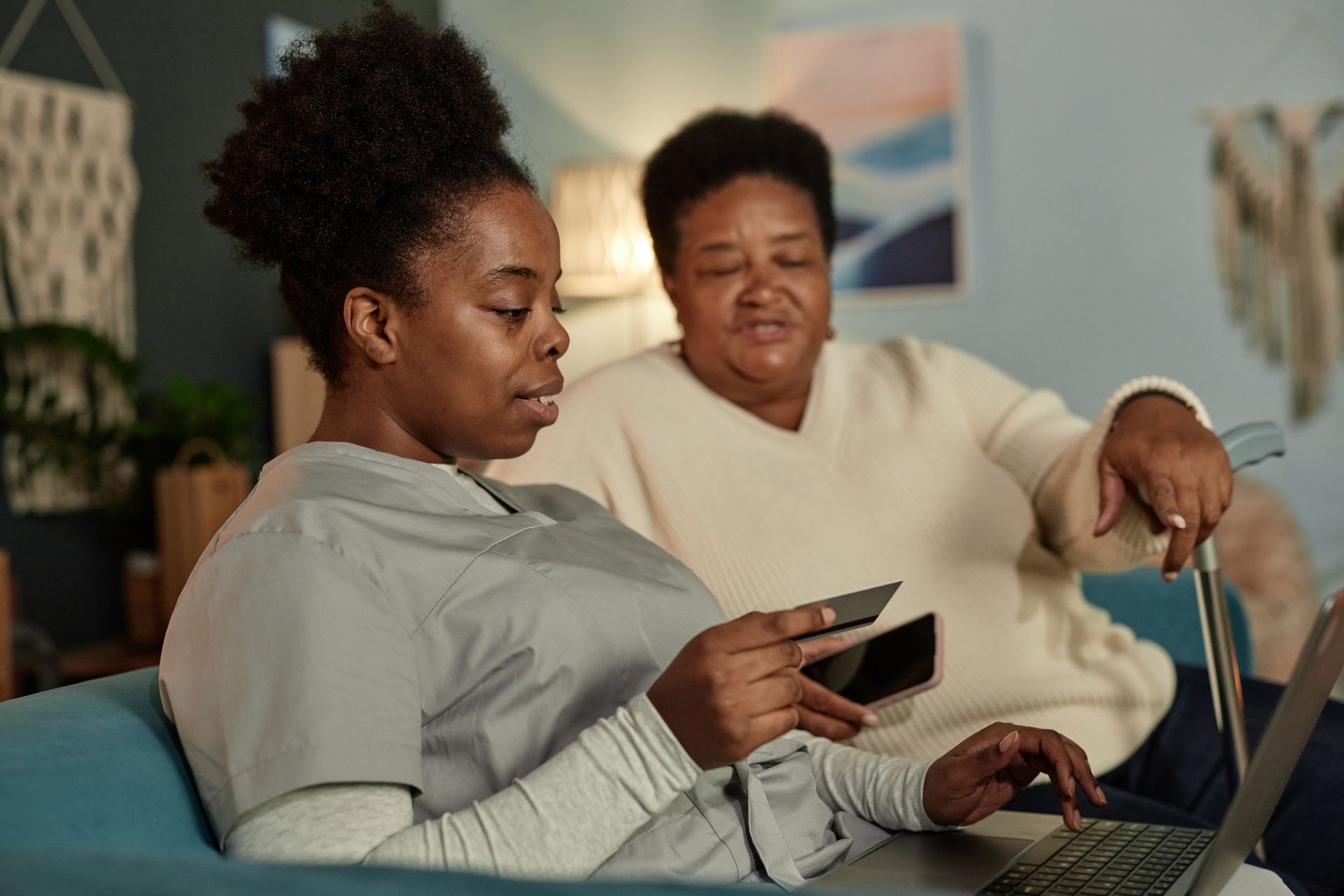 Two women are sitting on a couch using a laptop and a cell phone.