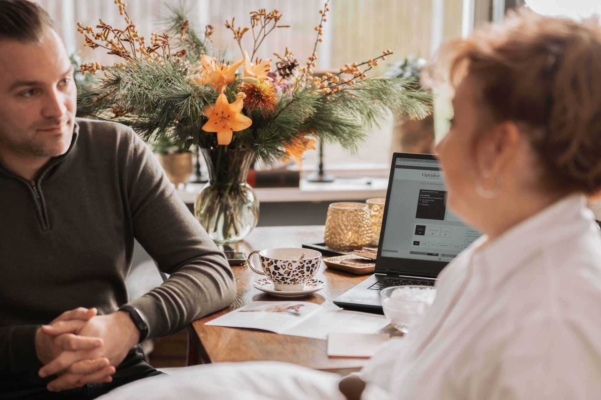 Een man en een vrouw zitten aan een tafel met een laptop, bloemen en een kop koffie; de ​​man kijkt naar de vrouw.