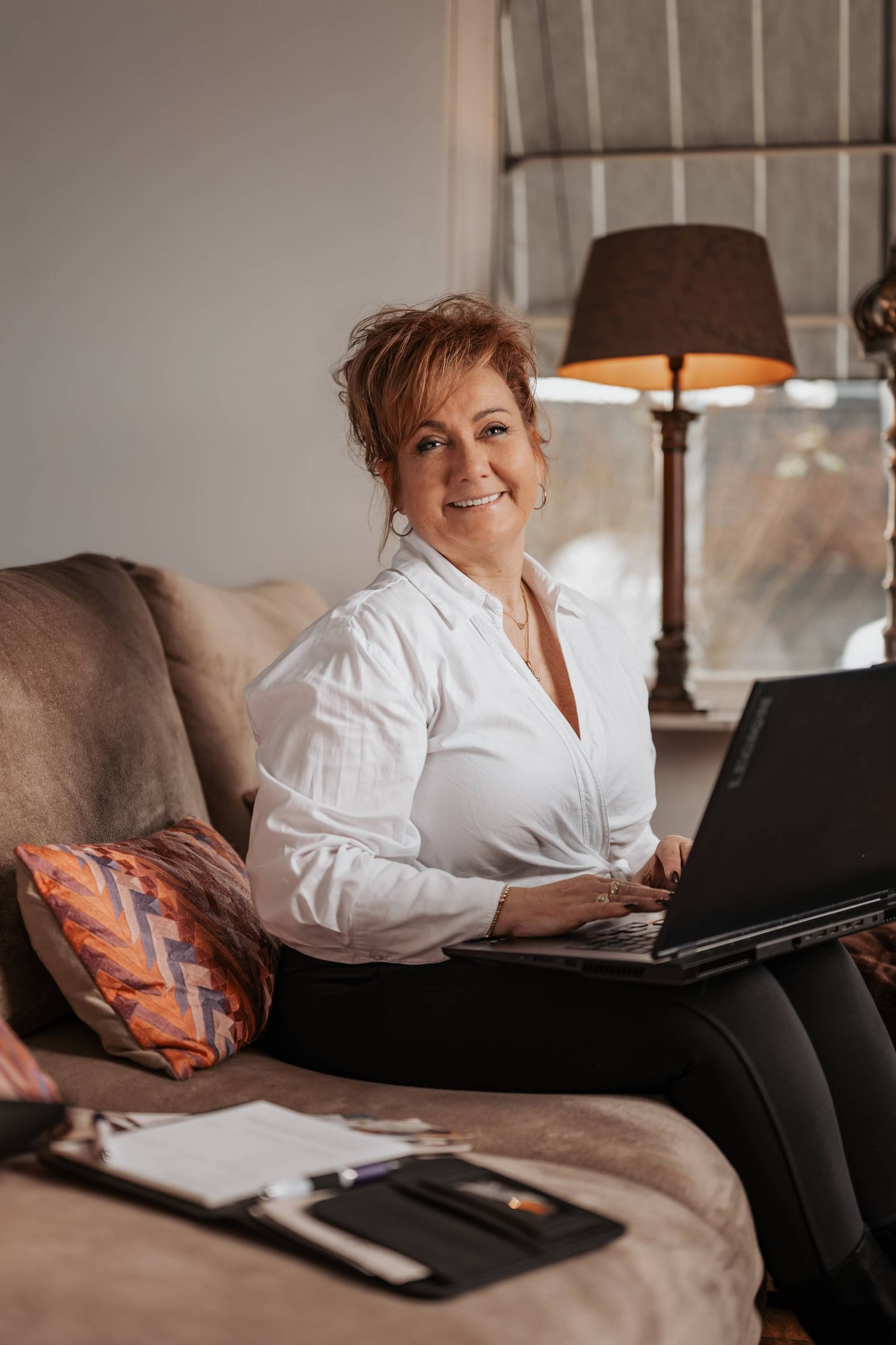 Vrouw zit op een bank, werkt met een laptop en lacht. Koffiekopje op de voorgrond, lamp en raam op de achtergrond.