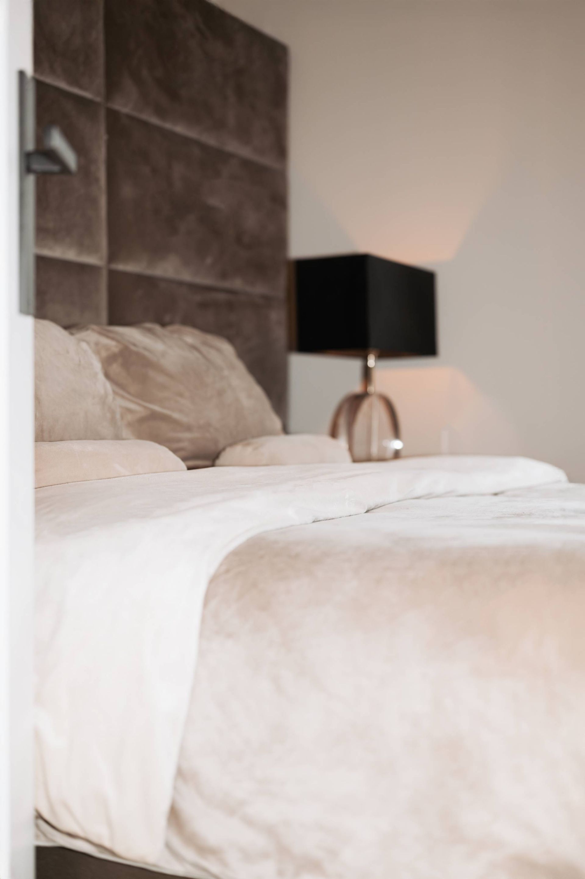 Bed met een gestoffeerd bruin hoofdeinde, beige beddengoed en een zwarte lampenkap.