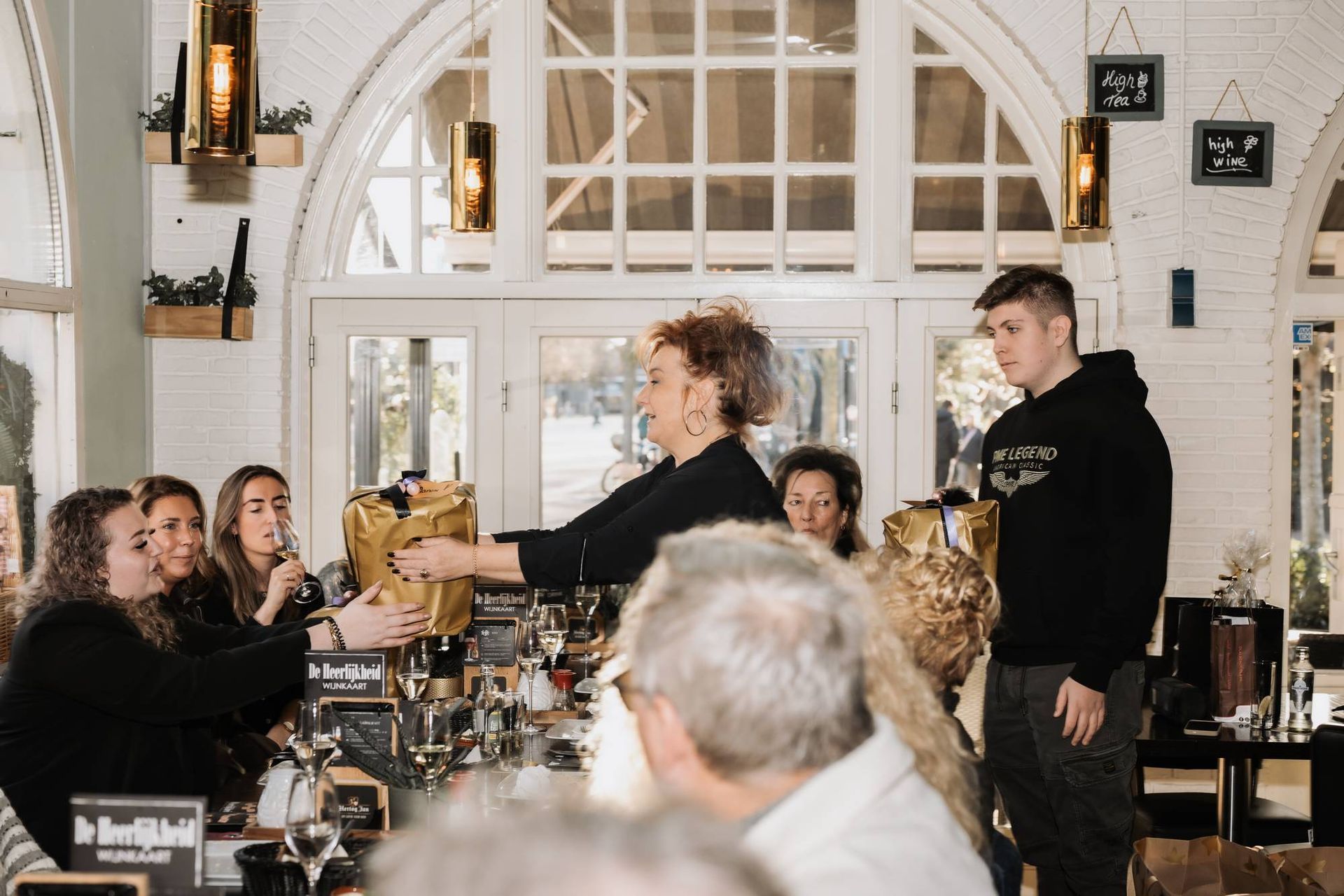 Mensen aan een tafel in een licht gebouw met boogvormige ramen; een vrouw overhandigt een gouden cadeautas aan iemand.