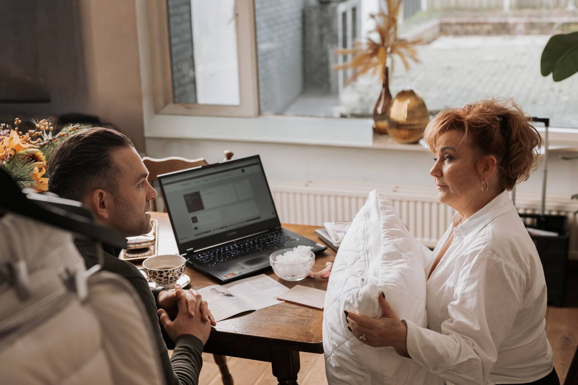 Vrouw met een wit kussen in haar handen praat met een man aan een tafel met een laptop en documenten.