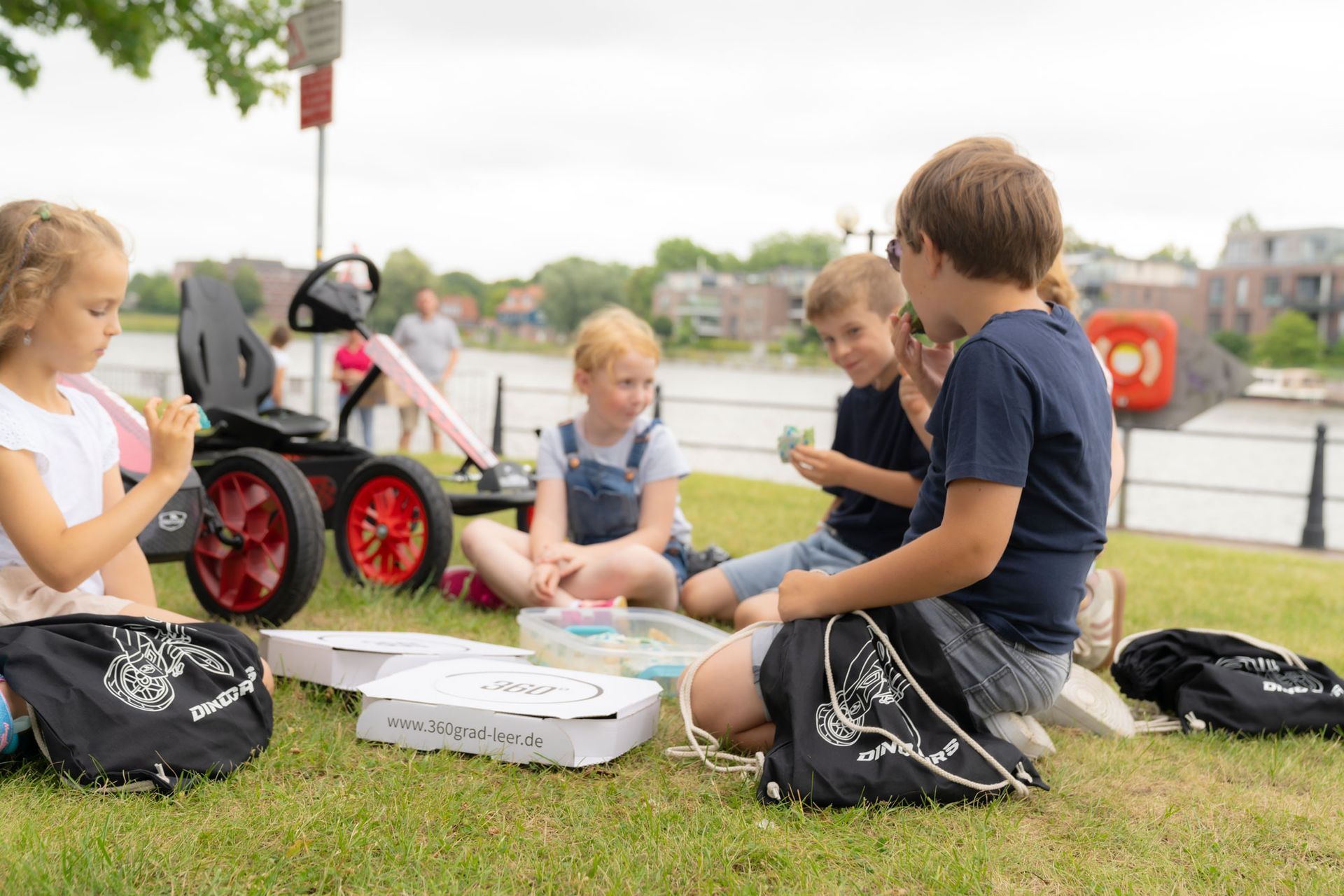 Kinder spielen auf einer Wiese in der Nähe eines Gewässers; im Hintergrund ist ein rotes Dino-Cars Go-Kart zu sehen.