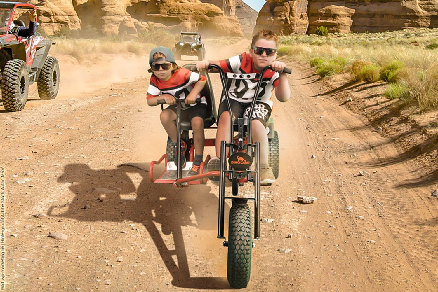 Zwei Jungen fahren mit einem umgebauten, dreirädrigen Motorrad auf einem unbefestigten Weg durch eine Wüstenlandschaft.