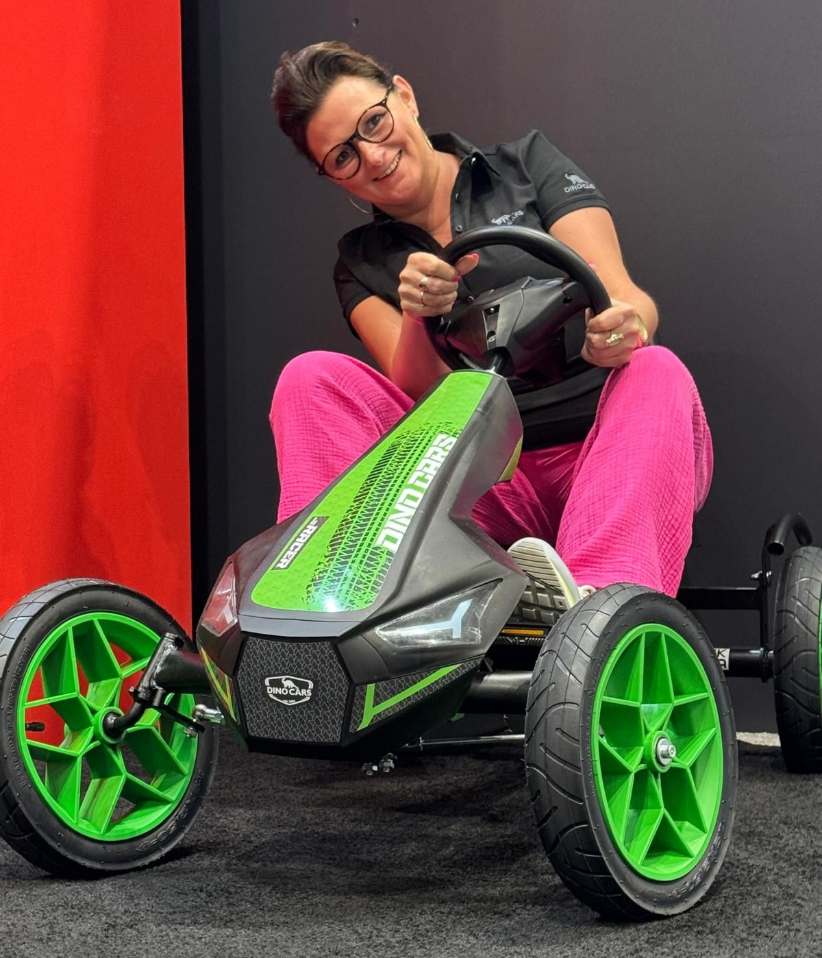 Frau sitzt in einem schwarz-grünen Go-Kart, die Hände am Lenkrad, trägt eine rosa Hose und lächelt.