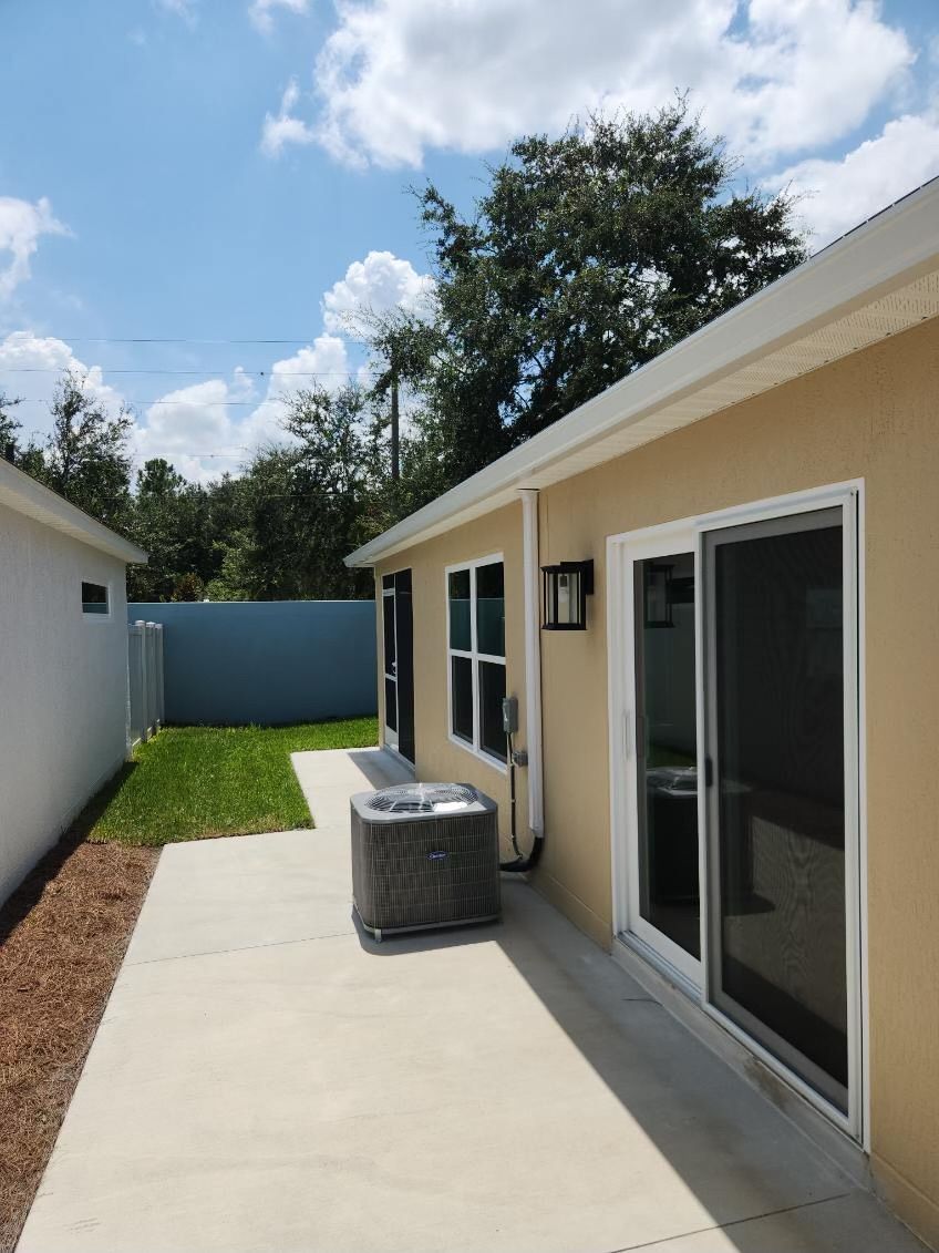 Backyard patio with tan house, sliding glass door, A/C unit, concrete path, and green lawn under a blue sky.
