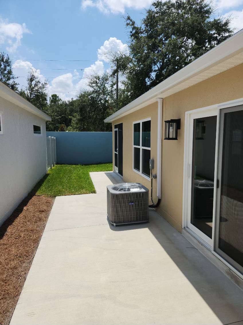 Backyard with concrete path, grass, and buildings in beige and white. Air conditioning unit visible.