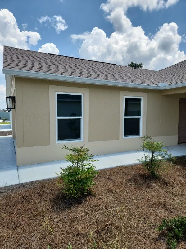 Tan house with two windows, brown roof, cloudy sky. Landscaping with small bushes.