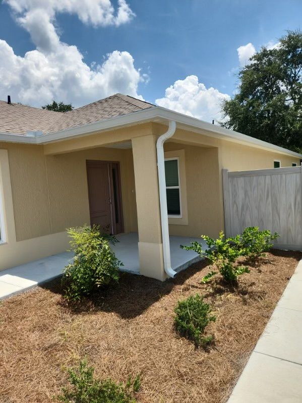 Tan house with white trim, porch, and garden. Blue sky with clouds in the background.
