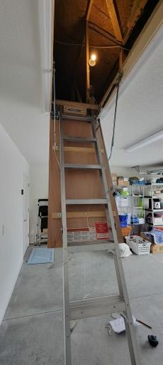 A pull-down attic ladder extended into a garage. The ladder is open, leading up to a dark attic space.
