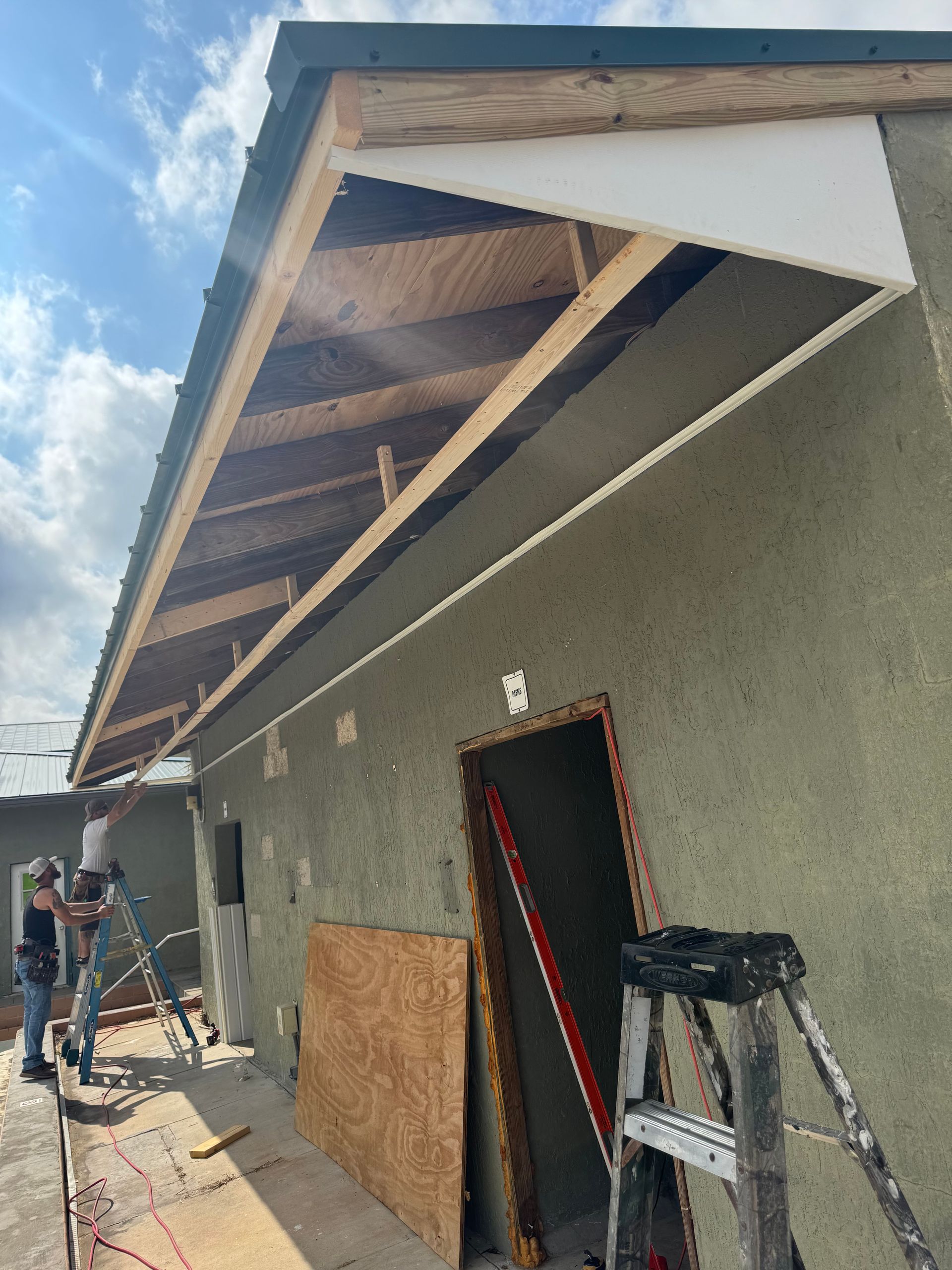 Construction worker on ladder, stucco building with exposed wood framing, blue sky.