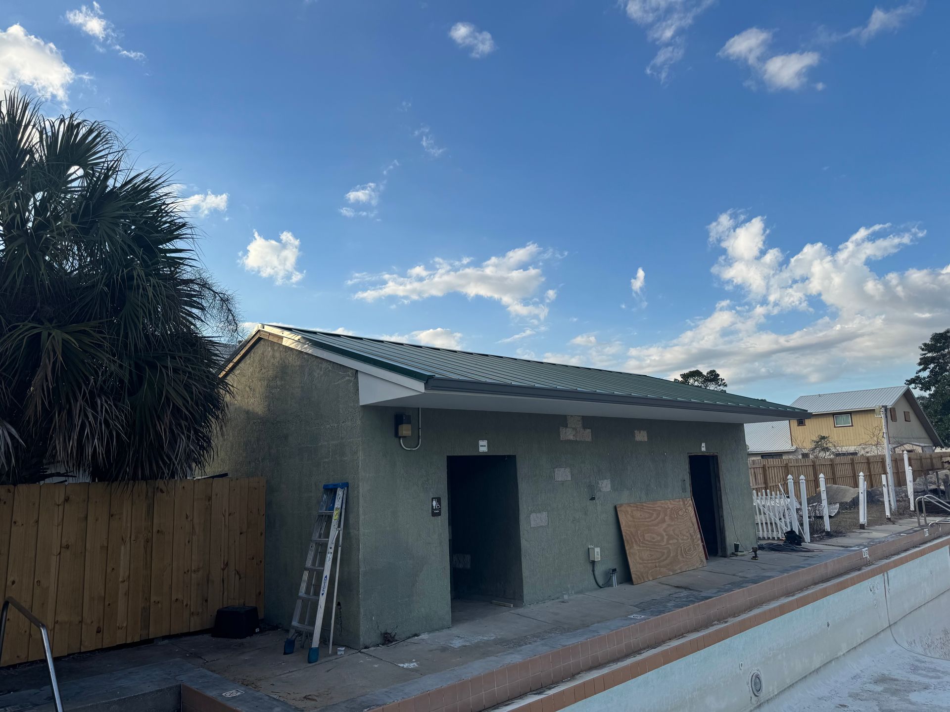 Pool house under construction with green stucco walls and a metal roof, next to a partially built pool.