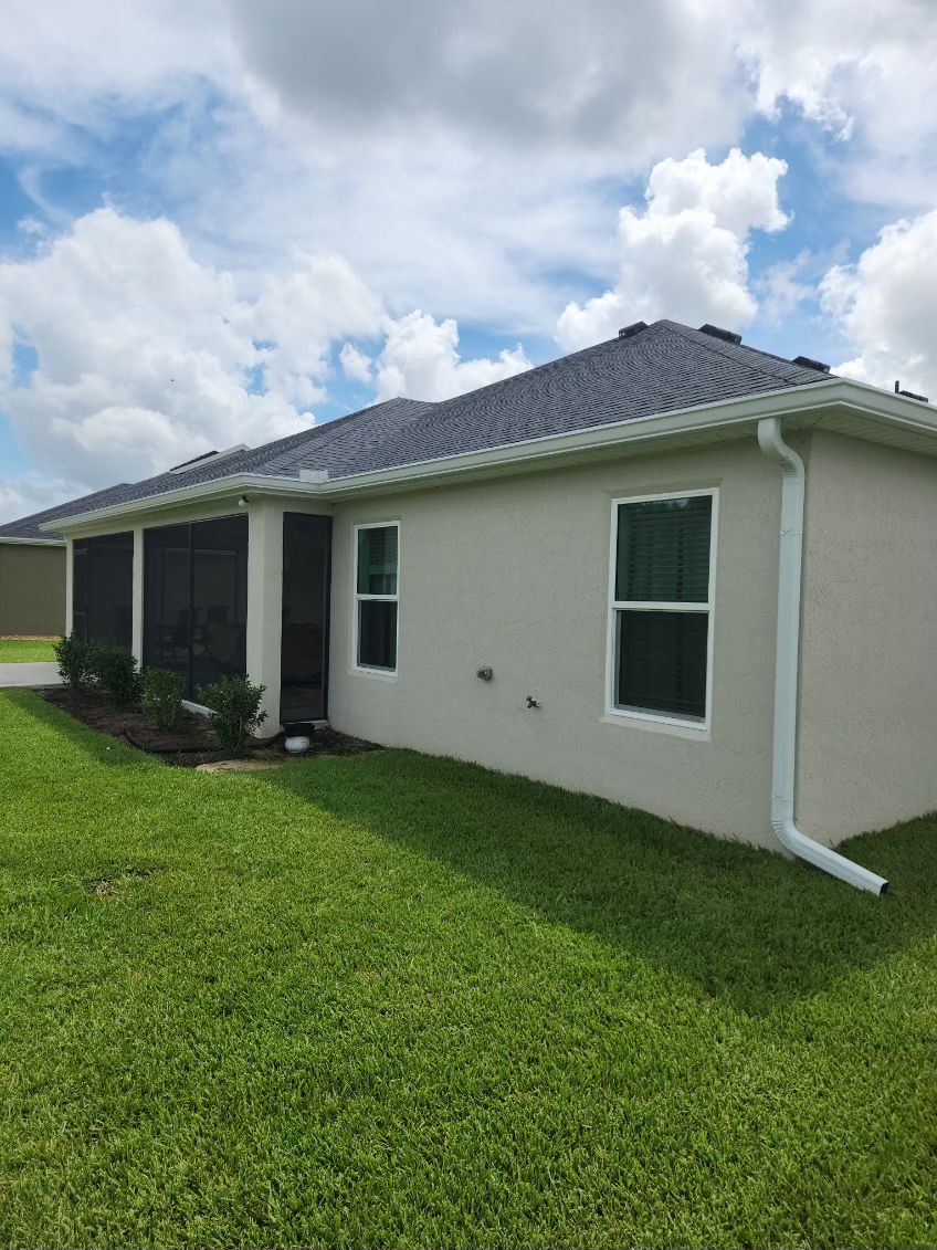 Beige house with dark roof, white trim, green grass, and blue sky with clouds.