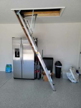 Attic access ladder deployed from ceiling in a garage. Stainless steel fridge, trash can, and clutter are present.